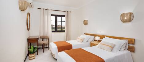 Quarto de hotel com duas camas, uma mesinha, uma cadeira, um vaso de planta e uma janela com cortinas.