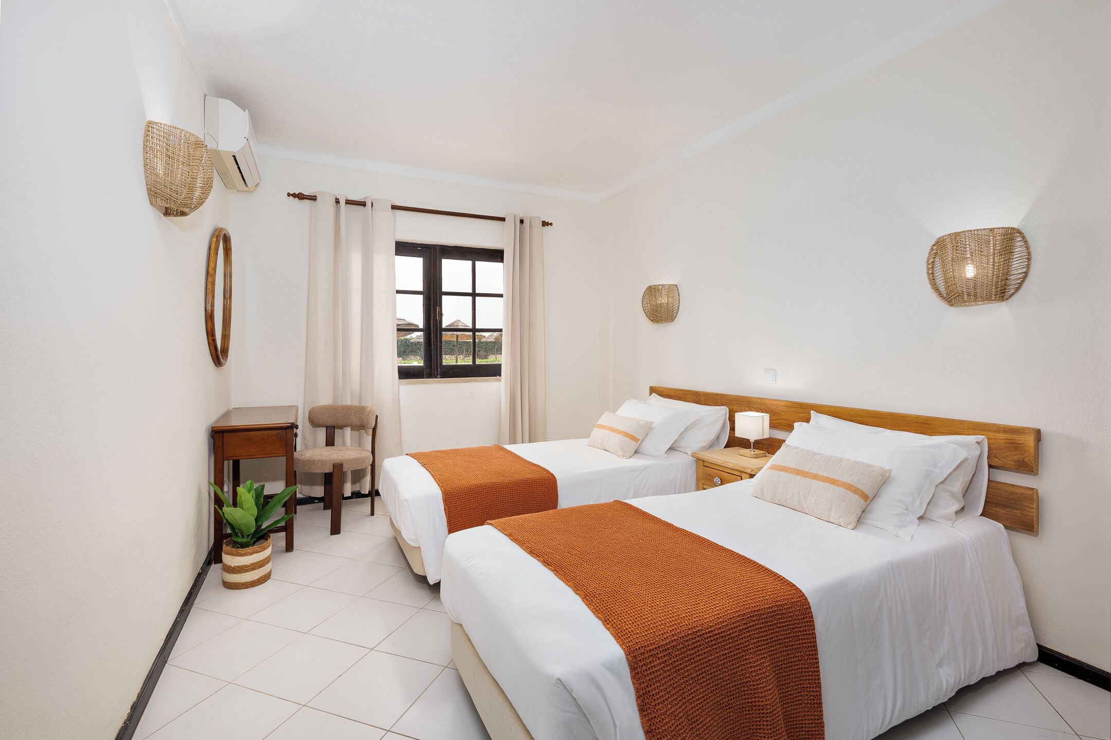 Une chambre d’hôtel avec deux lits, une petite table, une chaise, une plante en pot et une fenêtre avec des rideaux.