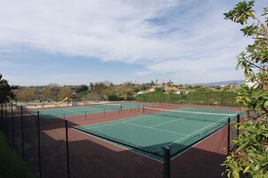 An outdoor tennis court with a net, surrounded by a fence, and a playground in the background.