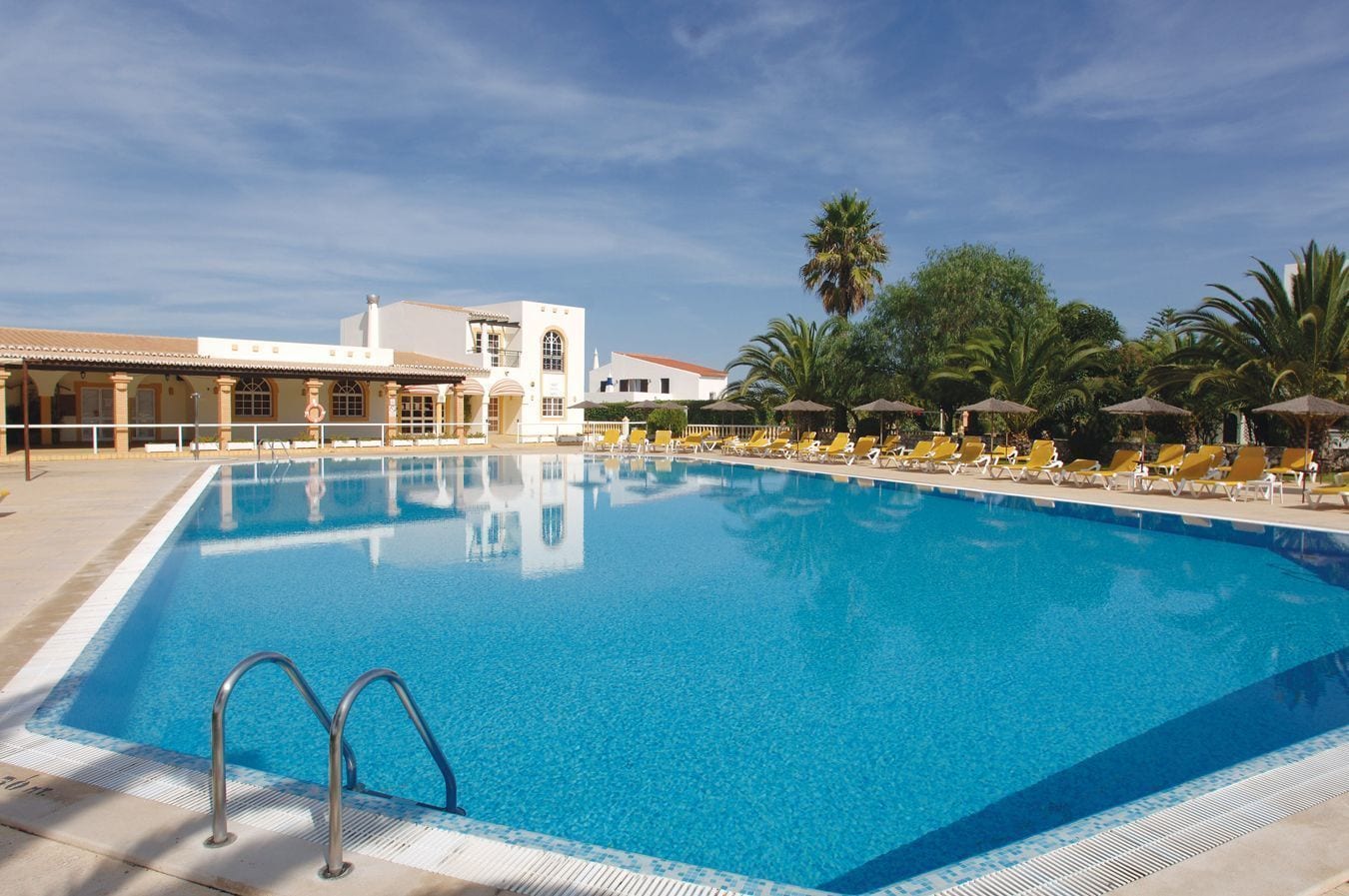 Ein rechteckiger Swimmingpool mit klarem, blauem Wasser, umgeben von Liegestühlen und Sonnenschirmen, mit einem Gebäude im Hintergrund.