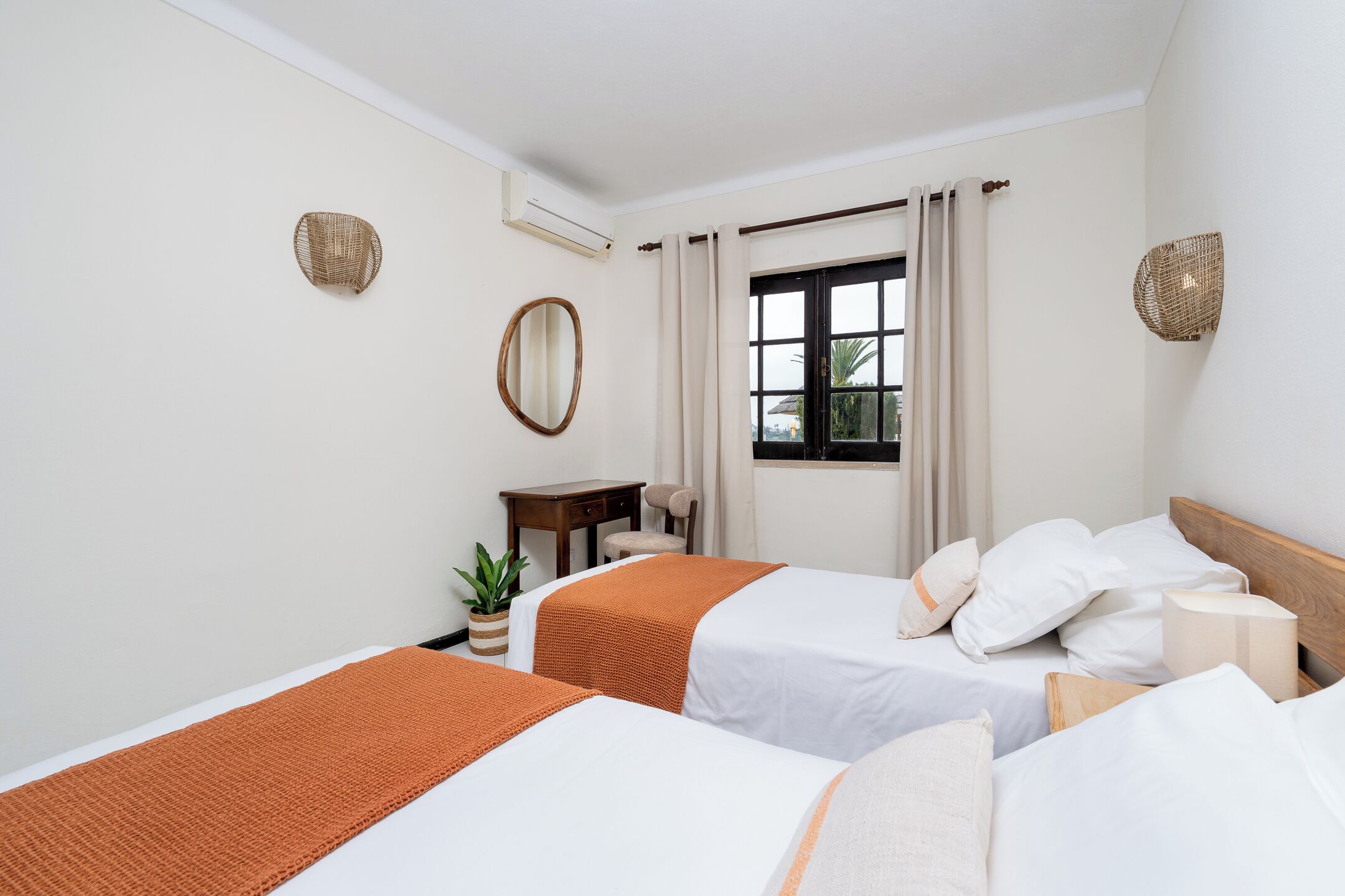 Habitación de hotel con dos camas, una mesita, una silla, una planta en maceta y una ventana con cortinas.