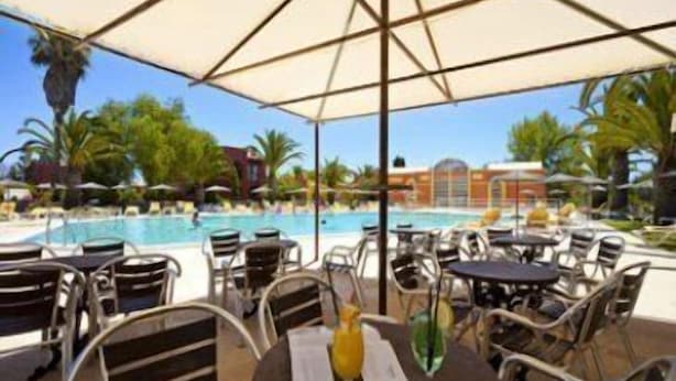 Zona de piscina al aire libre con mesas y sillas bajo sombrillas, con vistas a una piscina.