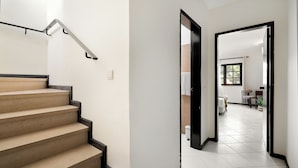 Una escalera con barandilla, un acceso a una habitación con ventana, y suelo de baldosas.