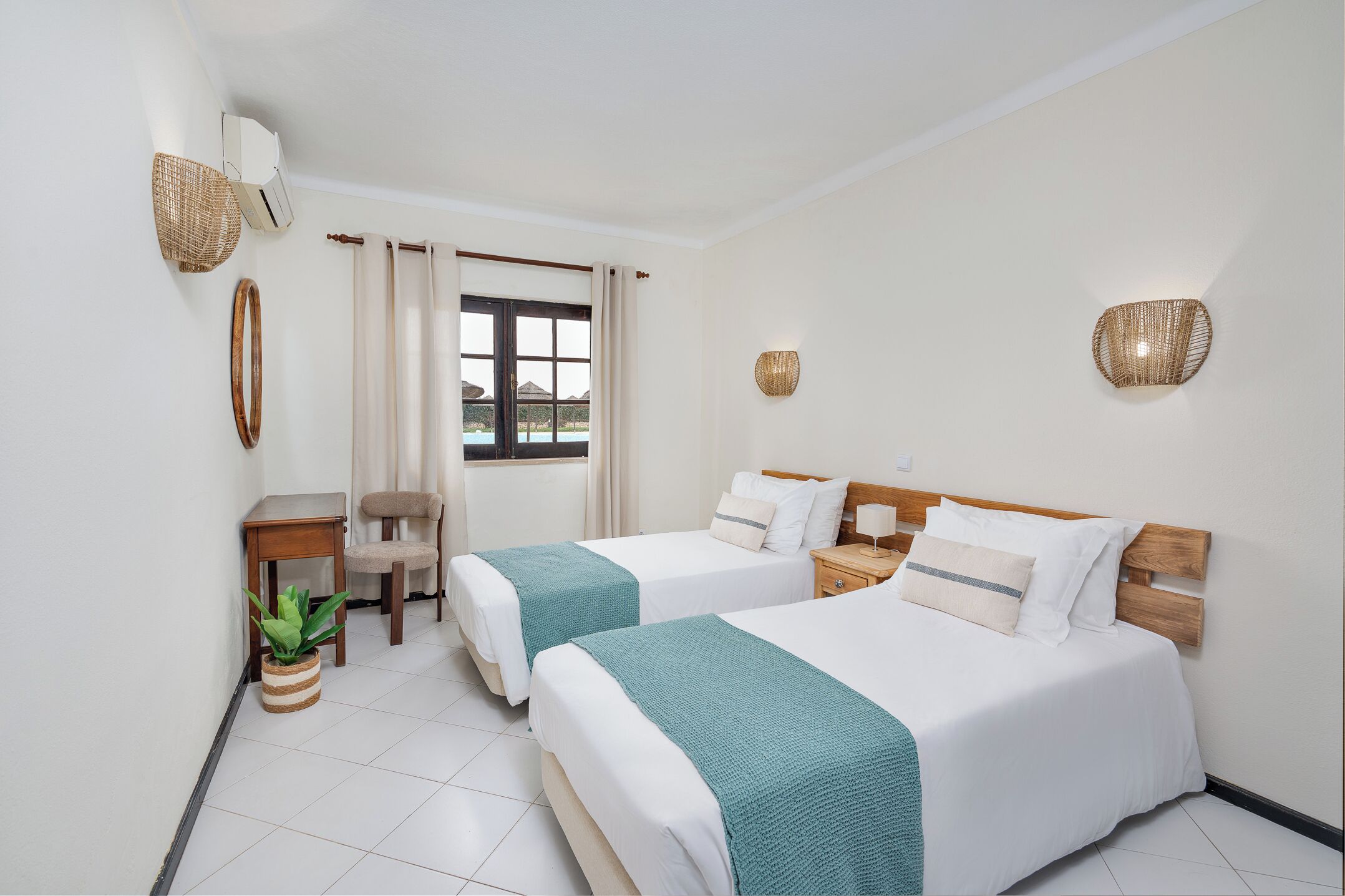 Une chambre d’hôtel avec deux lits, une petite table, une chaise, une plante en pot et une fenêtre avec des rideaux.