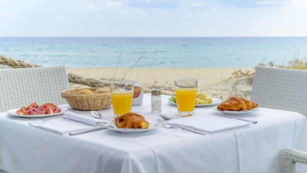 Una tavola imbandita con prodotti per la colazione, tra cui croissant, frutta e succo d'arancia, con vista sulla spiaggia.