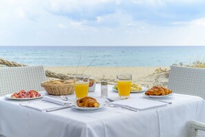 Una mesa dispuesta con alimentos para el desayuno, como cruasanes, fruta y jugo de naranja, con vista a la playa.