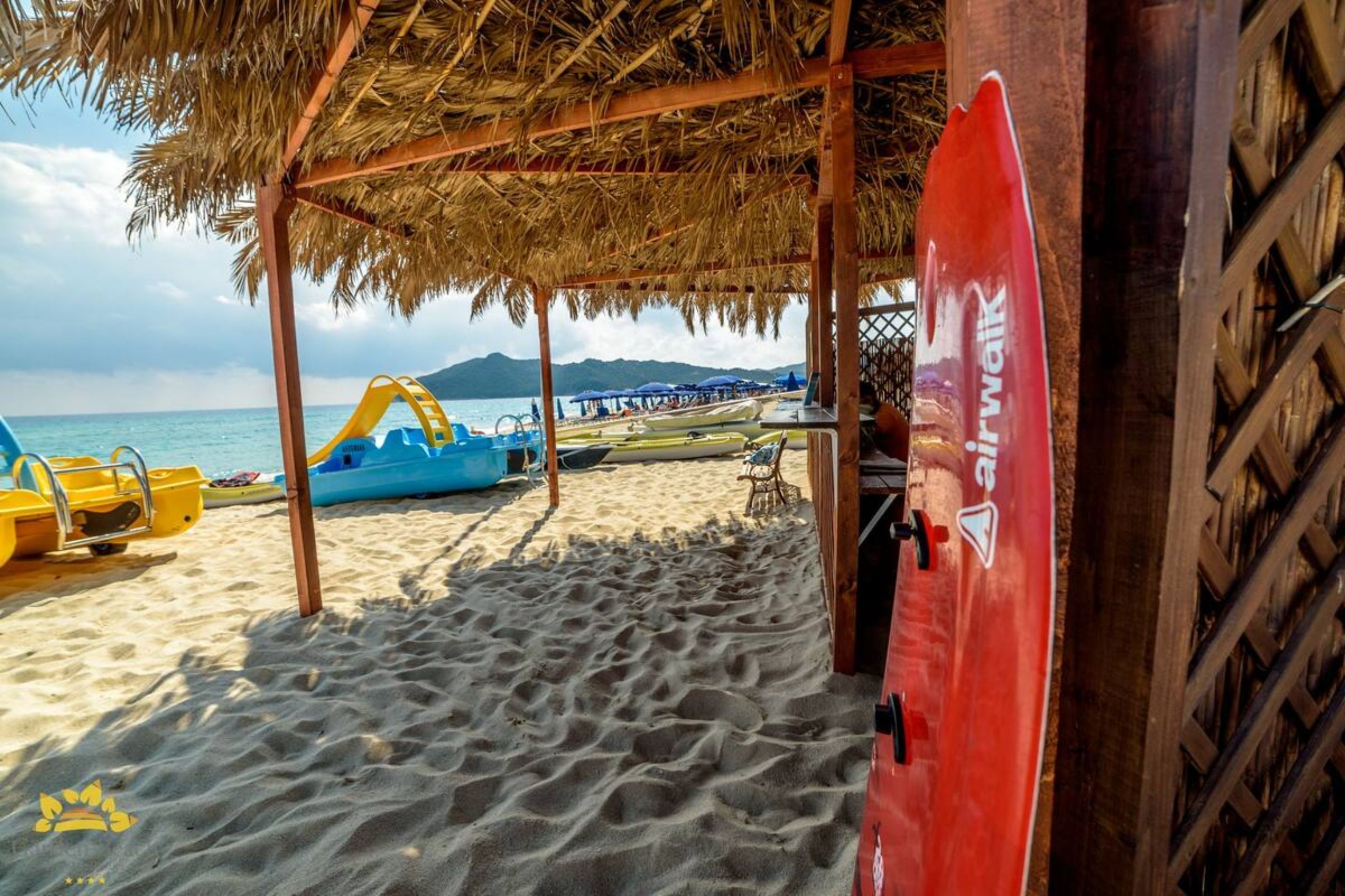 A beach area with a thatched roof structure, a red surfboard, and beach equipment.