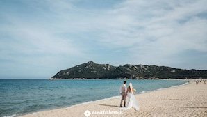 Una spiaggia con una coppia che cammina sulla riva, una montagna sullo sfondo e un cielo sereno.