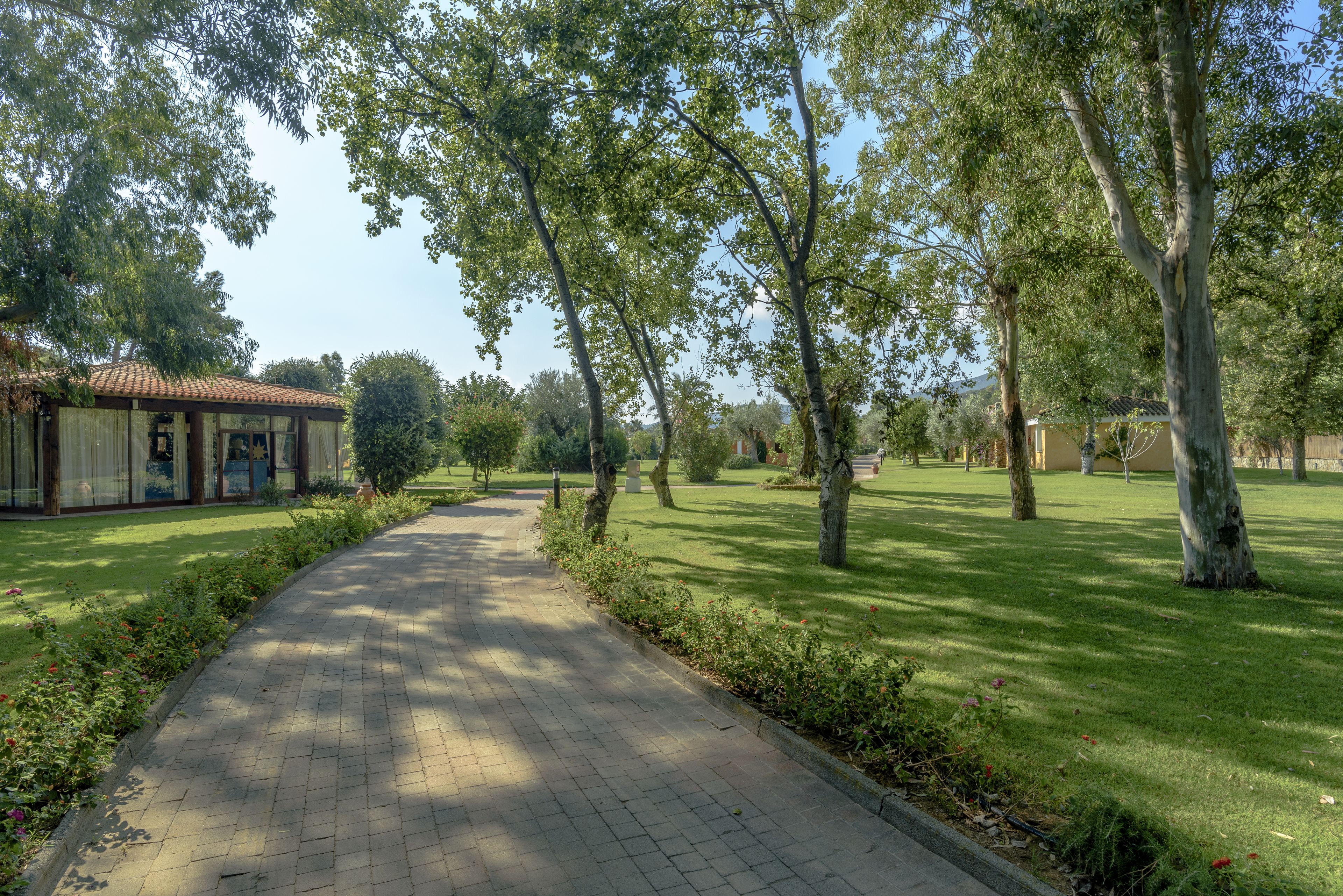 Un chemin pavé, aménagé dans un espace verdoyant avec des arbres et un bâtiment doté d’un toit recouvert de tuiles.