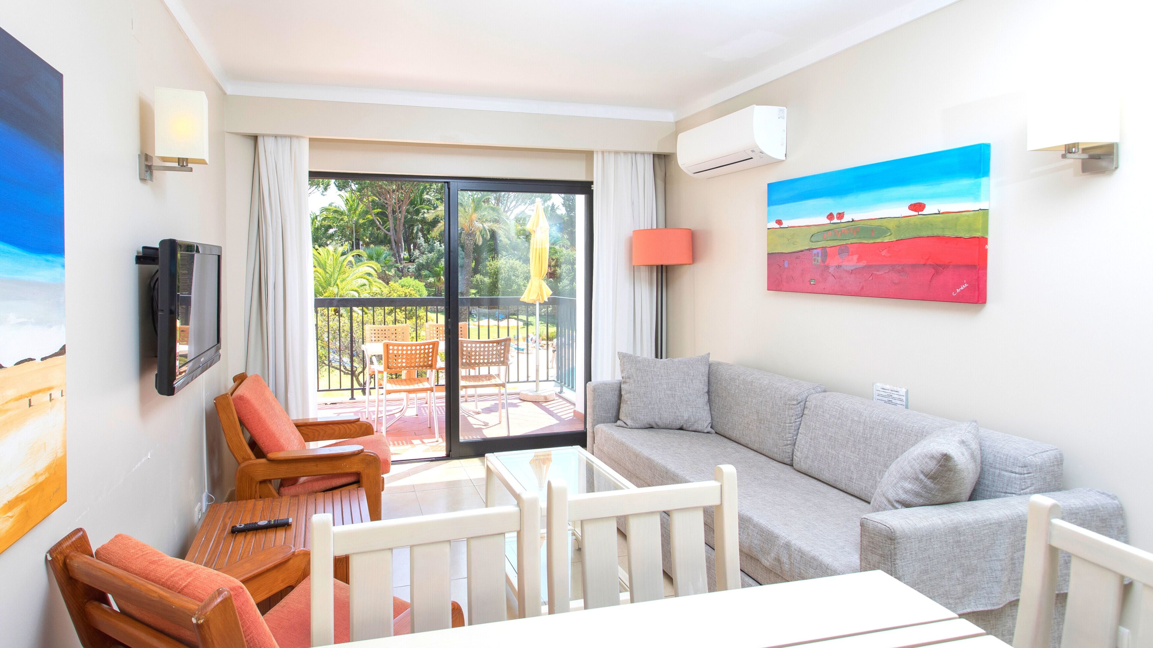 Uma sala de estar com um sofá cinzento, cadeiras de madeira, uma mesa de vidro, uma televisão e um quadro na parede.
