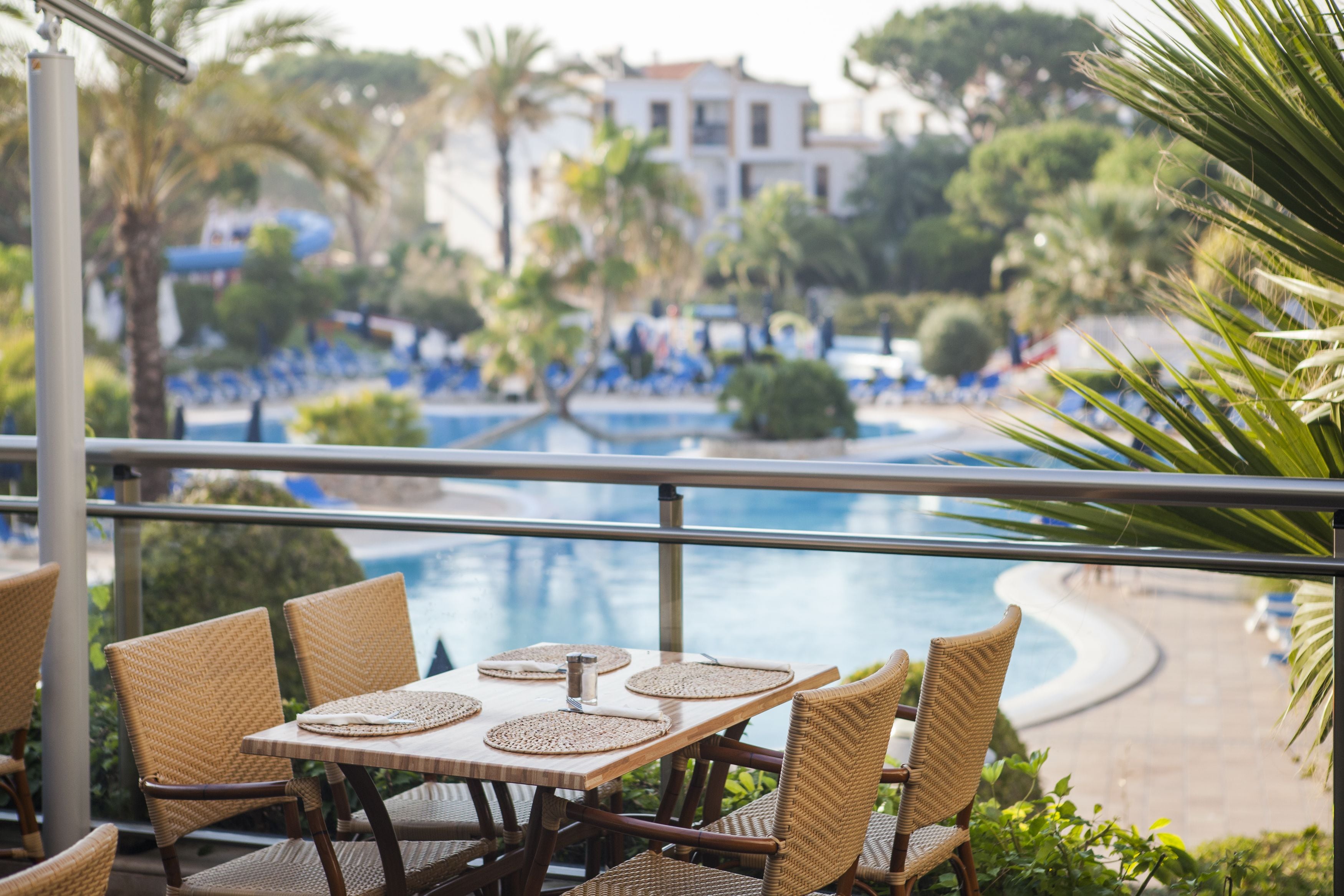 Un área de comedor junto a la piscina con sillas y mesas de mimbre, con vista a una piscina y una zona de resort con palmeras y un edificio al fondo.