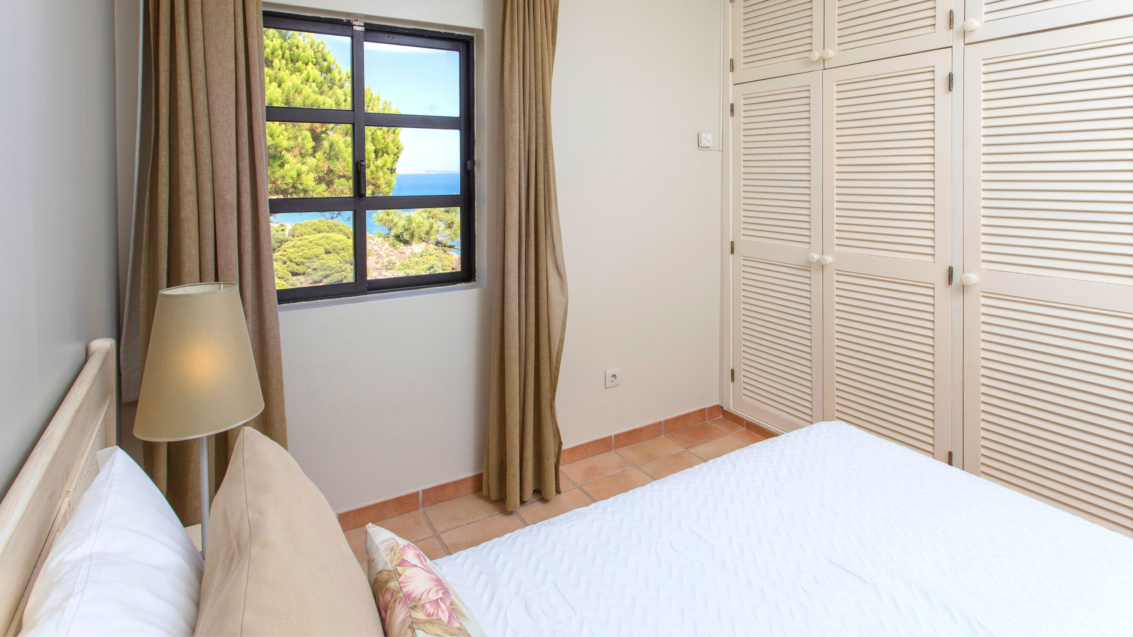 A bedroom with a bed, a window with curtains, a lamp, and a wardrobe.