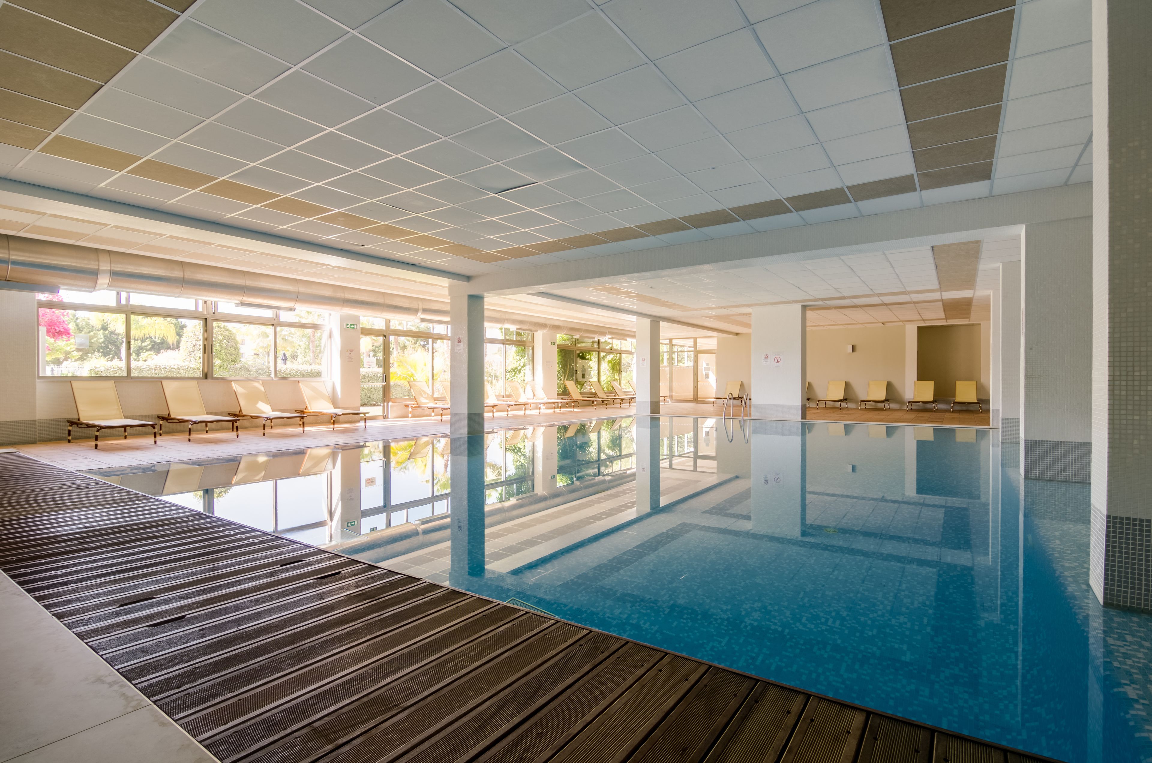 Una piscina interior con paredes de vidrio transparente, terraza de madera y zona de estar iluminada por el sol.