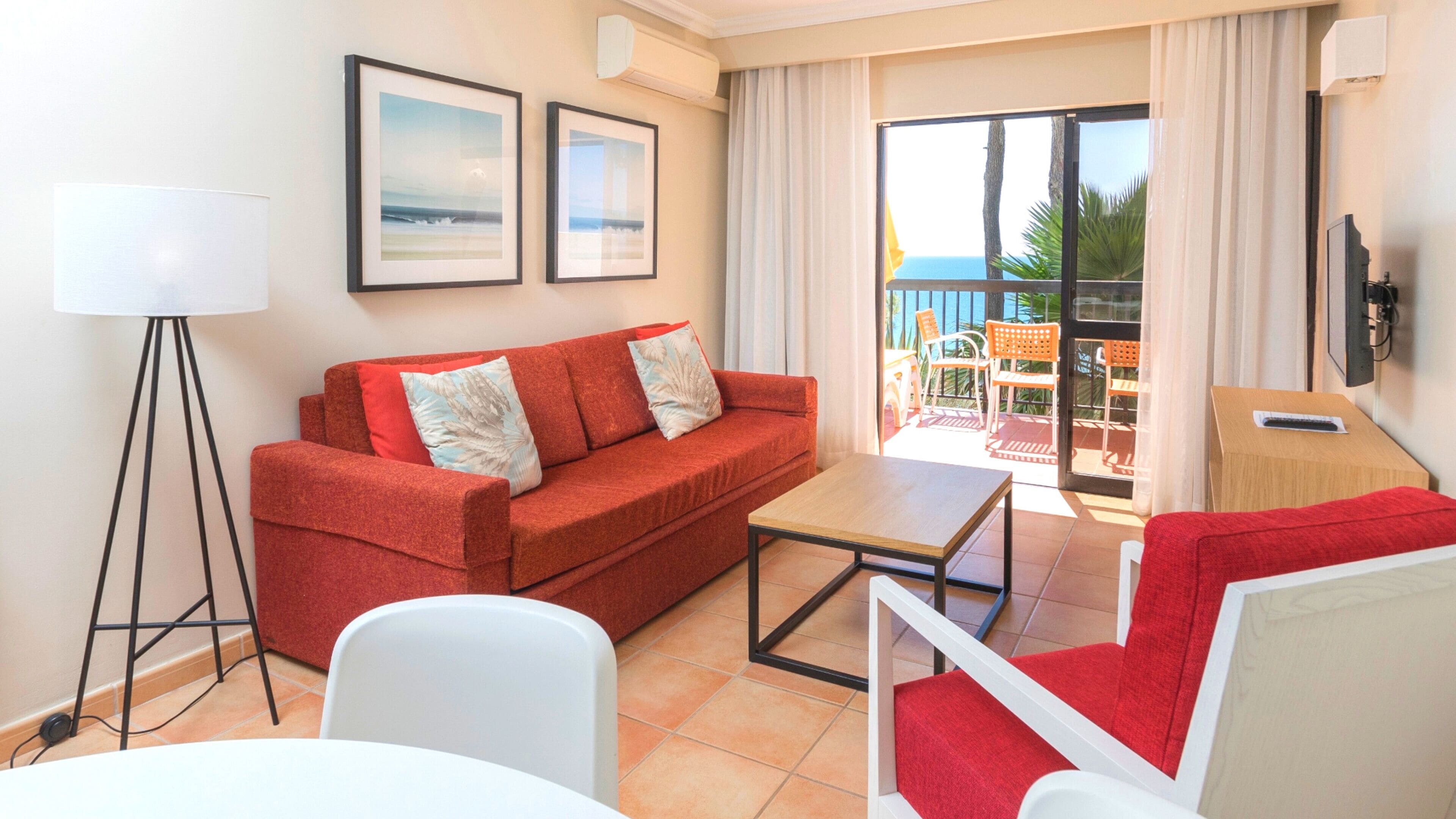 A modern living room with a red sofa, a white armchair, a small table, and a floor lamp. There are two framed pictures on the wall, a sliding glass door leading to a balcony with outdoor furniture, and a flat-screen TV mounted on the wall.