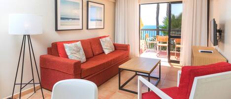 Un salon moderne avec un canapé rouge, un fauteuil blanc, une petite table et un lampadaire. Deux tableaux encadrés ornent le mur. Une porte-fenêtre coulissante donne sur un balcon aménagé avec du mobilier d’extérieur, et un téléviseur à écran plat est fixé au mur.