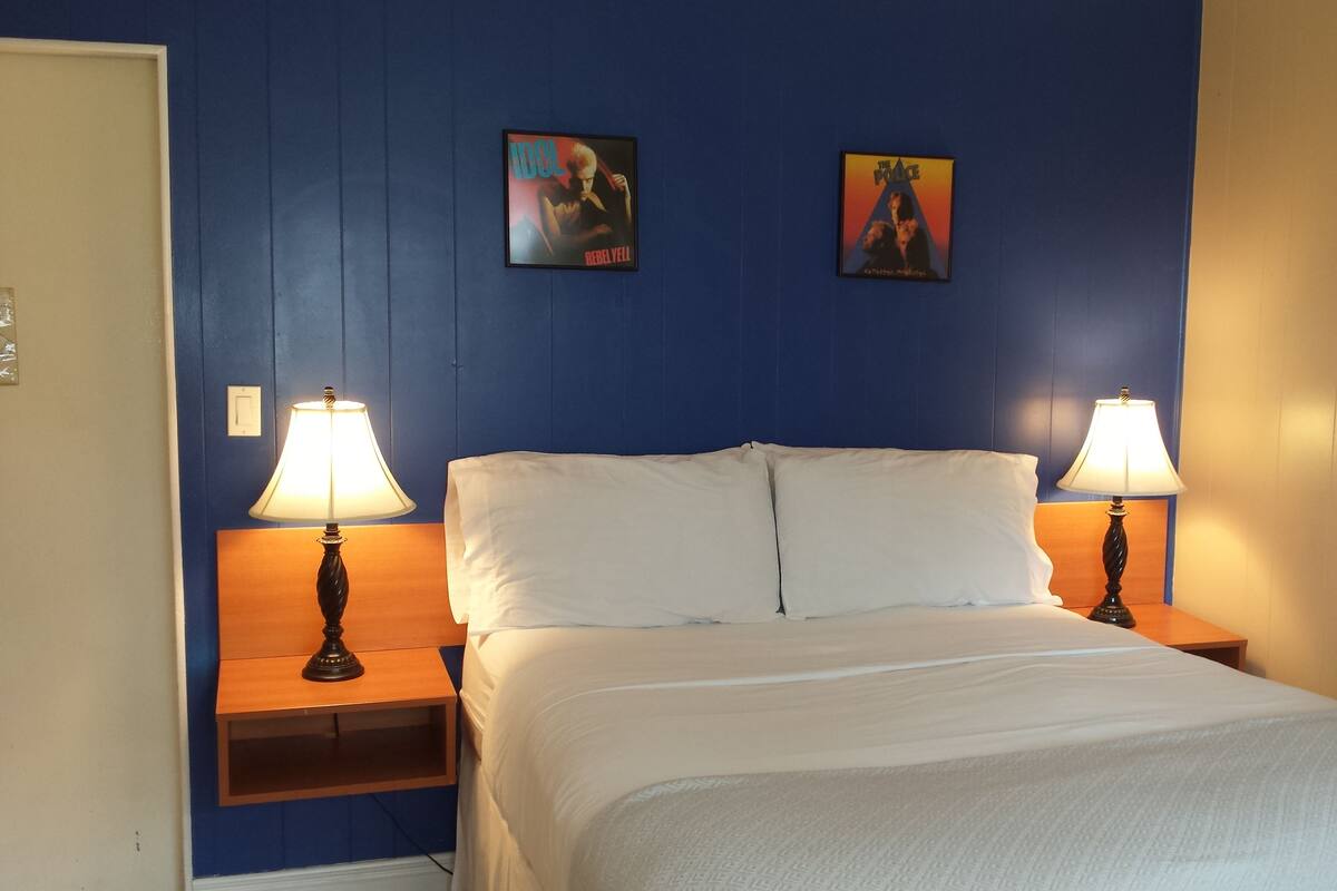 A hotel room with a bed, two bedside lamps, and two posters on the wall.