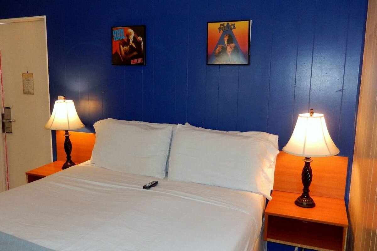 A hotel room with a bed, two bedside lamps, a remote control, and two posters on the wall.