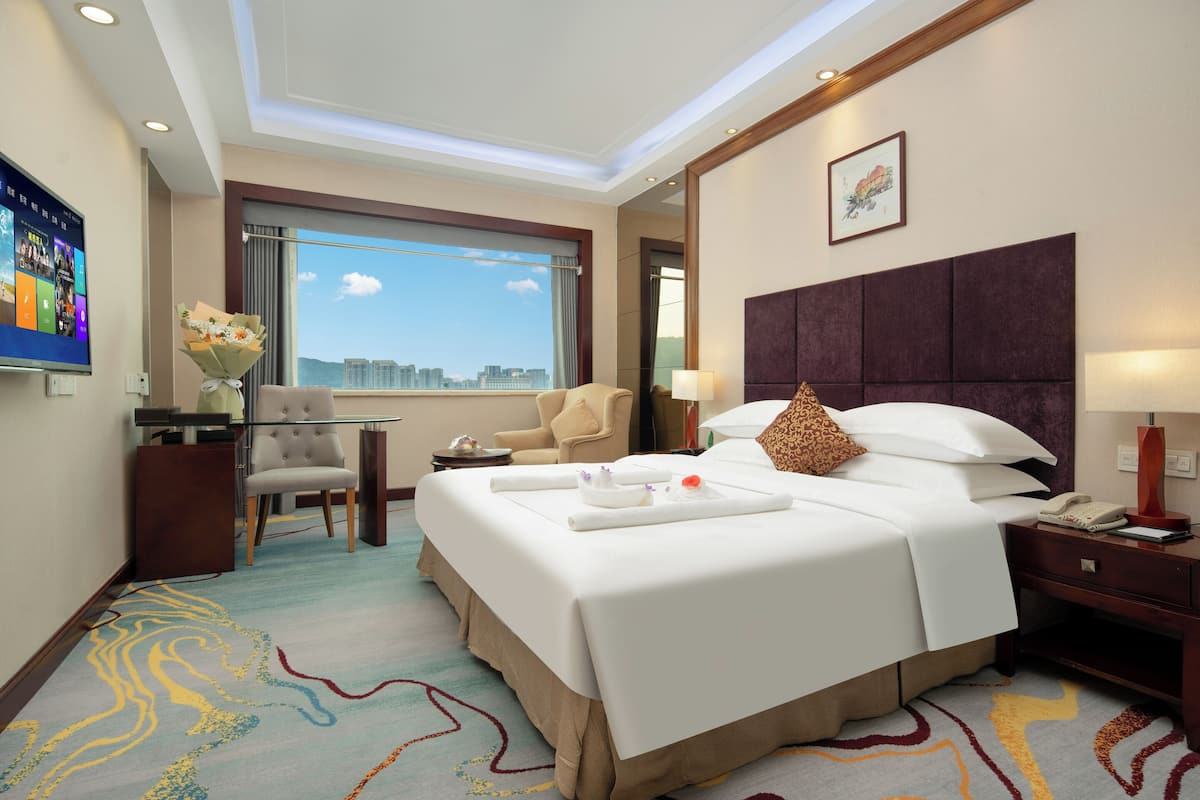 Ein Hotelzimmer mit einem großen Bett, einem Schreibtisch, einem Sessel, einem Fernseher und einem Fenster mit Blick auf die Stadt.