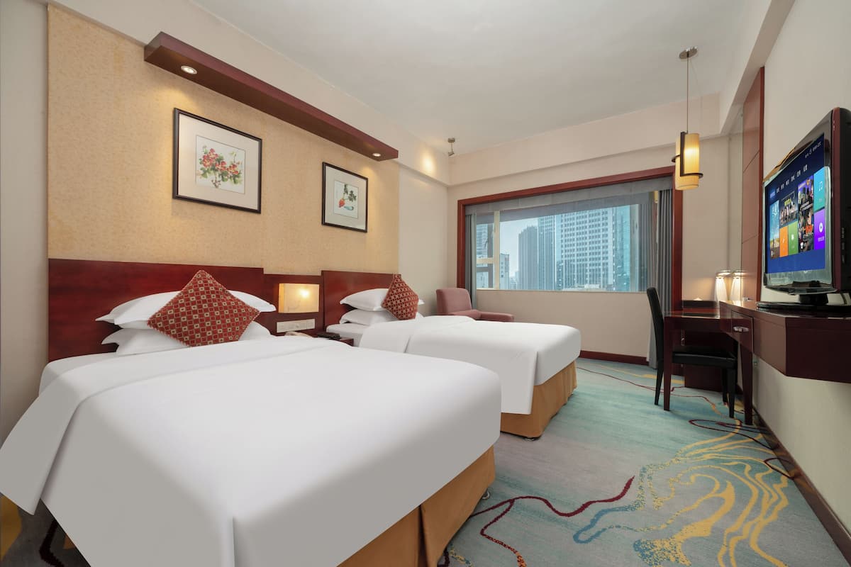 Ein Hotelzimmer mit zwei Betten, einem Schreibtisch mit Computer, einem Fernseher und einem Fenster mit Blick auf die Stadt.