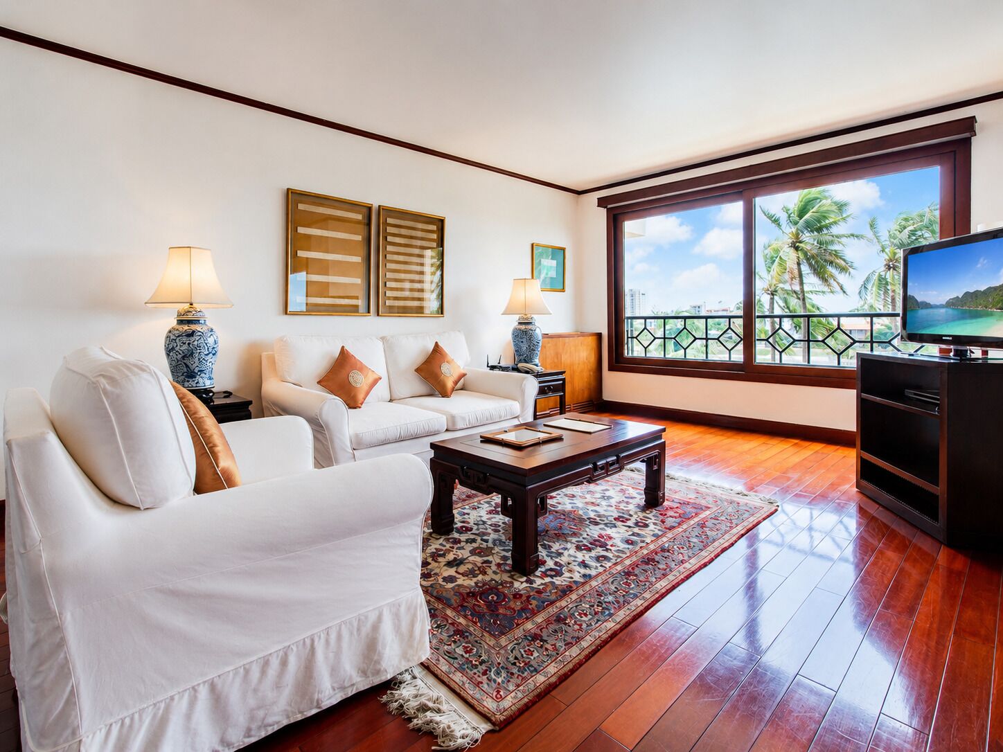 A living room with a sofa, coffee table, TV, and a view of palm trees through large windows.