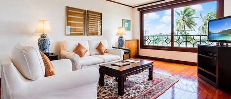 A living room with a sofa, coffee table, TV, and a view of palm trees through large windows.