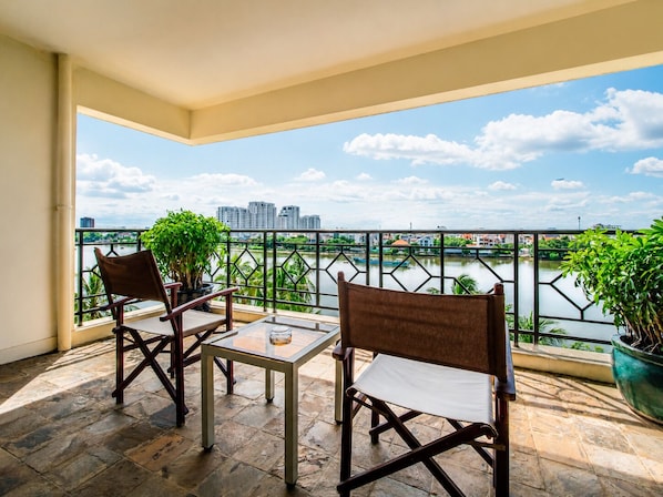 A balcony with two chairs and a table, overlooking a river with buildings in the distance.