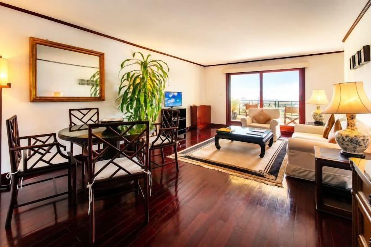 Una sala de estar con una mesa de comedor, sillas, un sofá, un televisor, una planta grande y vistas a través de una ventana.