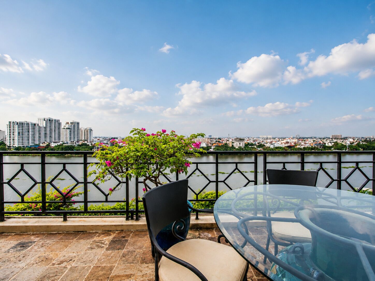 A terrace with a glass table and chairs, a view of a cityscape, and a railing with greenery.