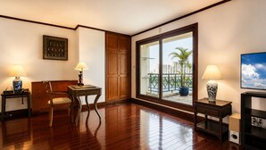 A room with a wooden floor, a desk with a lamp, a chair, a wardrobe, and a large window with a view of buildings and a palm tree.