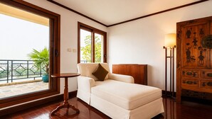 A room with a bed, a small round table, a large window with a view of greenery, and a traditional wooden cabinet.
