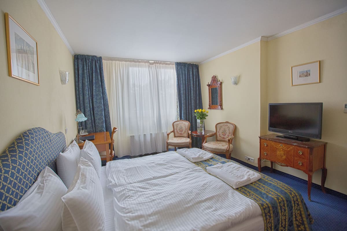 A hotel room with a large bed, two armchairs, a small table with a vase, a television on a wooden cabinet, and a window with curtains.