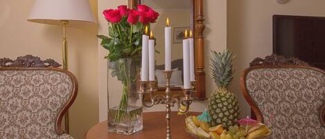 Una mesa redonda de madera con un candelabro, un florero con rosas rojas y un frutero. Dos sillones tapizados están colocados alrededor de la mesa.