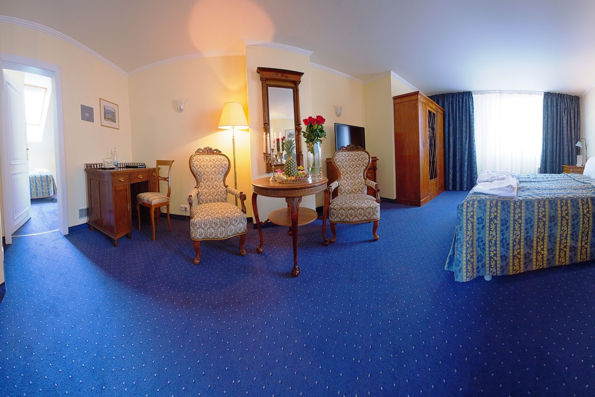 A hotel room with a bed, two chairs, a desk, a mirror, and a small table with a vase of flowers.