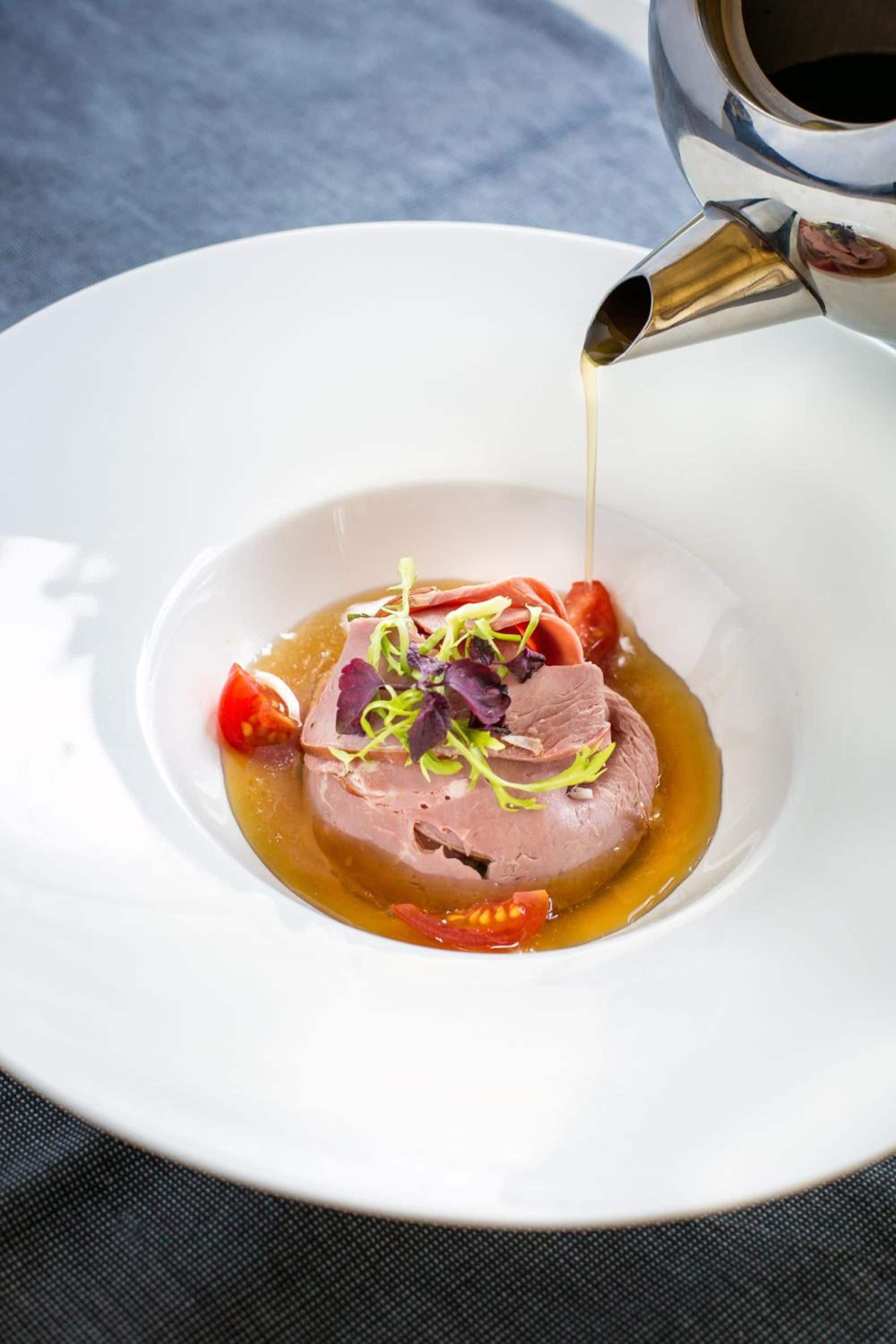 Un plato blanco con un guiso de carne, adornado con vegetales verdes y salsa vertiéndose sobre él.