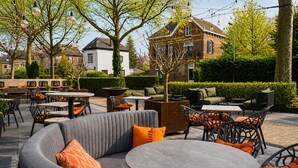 An outdoor seating area with round tables and chairs, surrounded by greenery and a building with a brick facade. - Hotel Hof Van Gelre by Flow (Lochem)