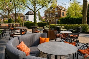 An outdoor seating area with round tables and chairs, surrounded by greenery and a building with a brick facade.