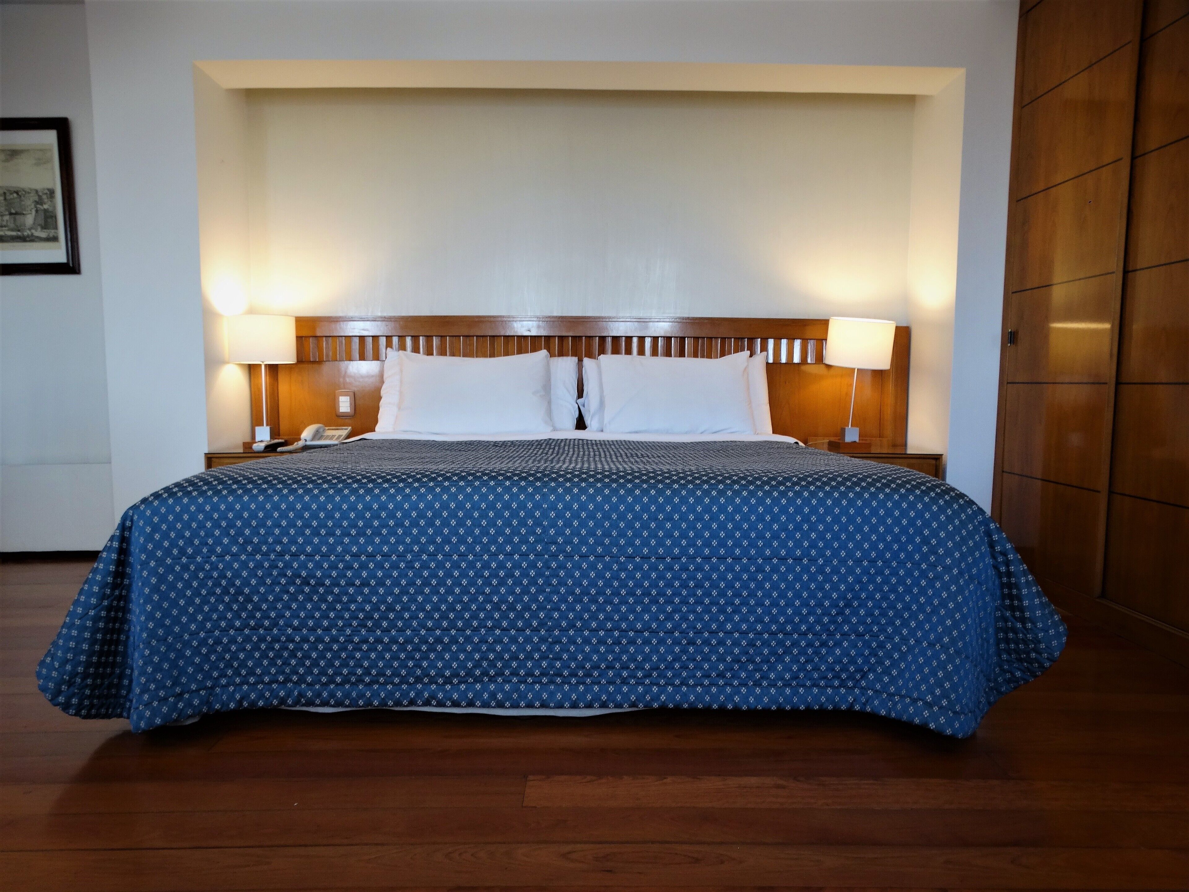A hotel room with a large bed, wooden headboard, two bedside lamps, and a framed picture on the wall.