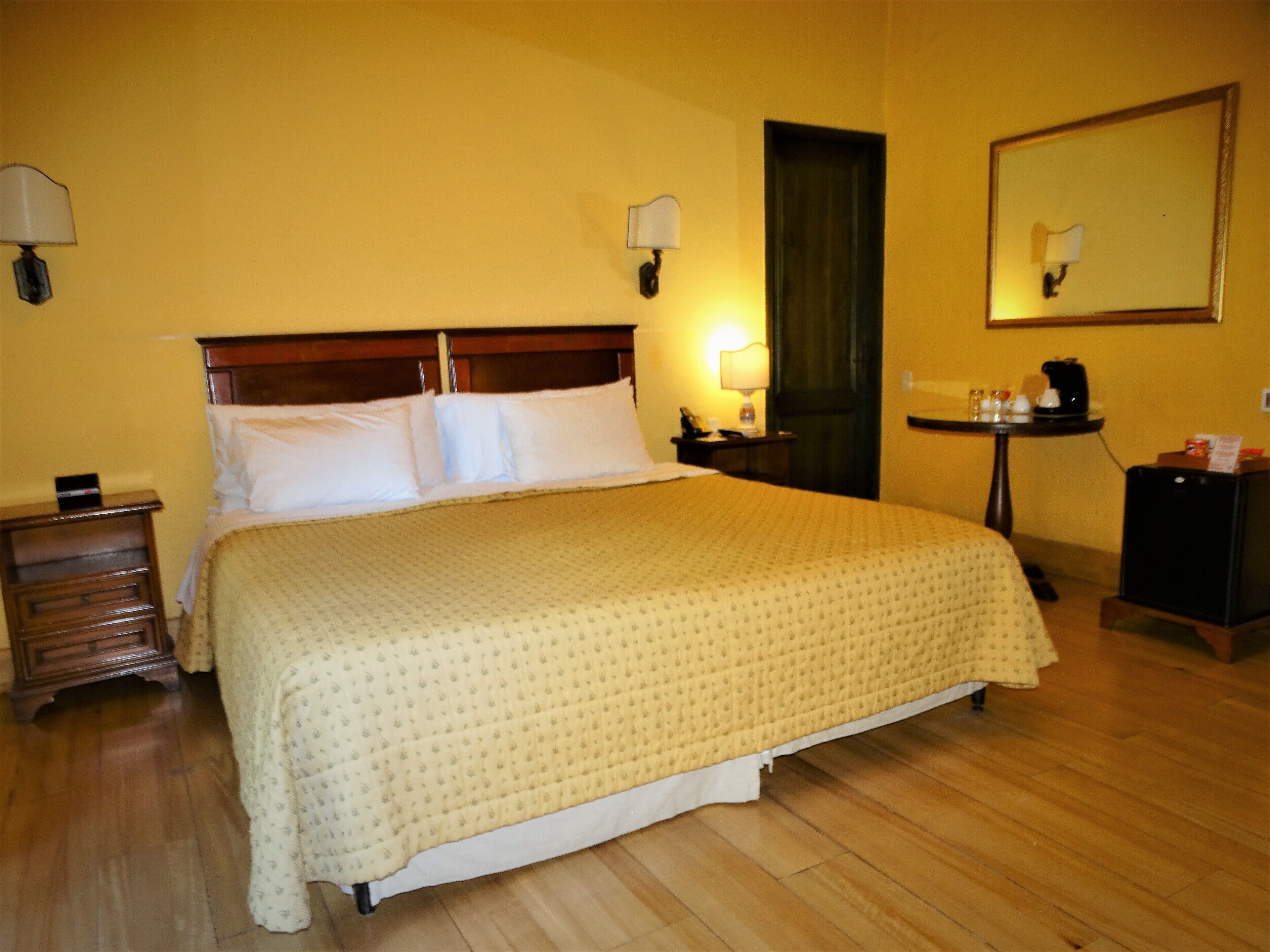 A hotel room with a large bed, wooden headboard, bedside tables with lamps, a small table with a coffee maker, and a mirror on the wall.