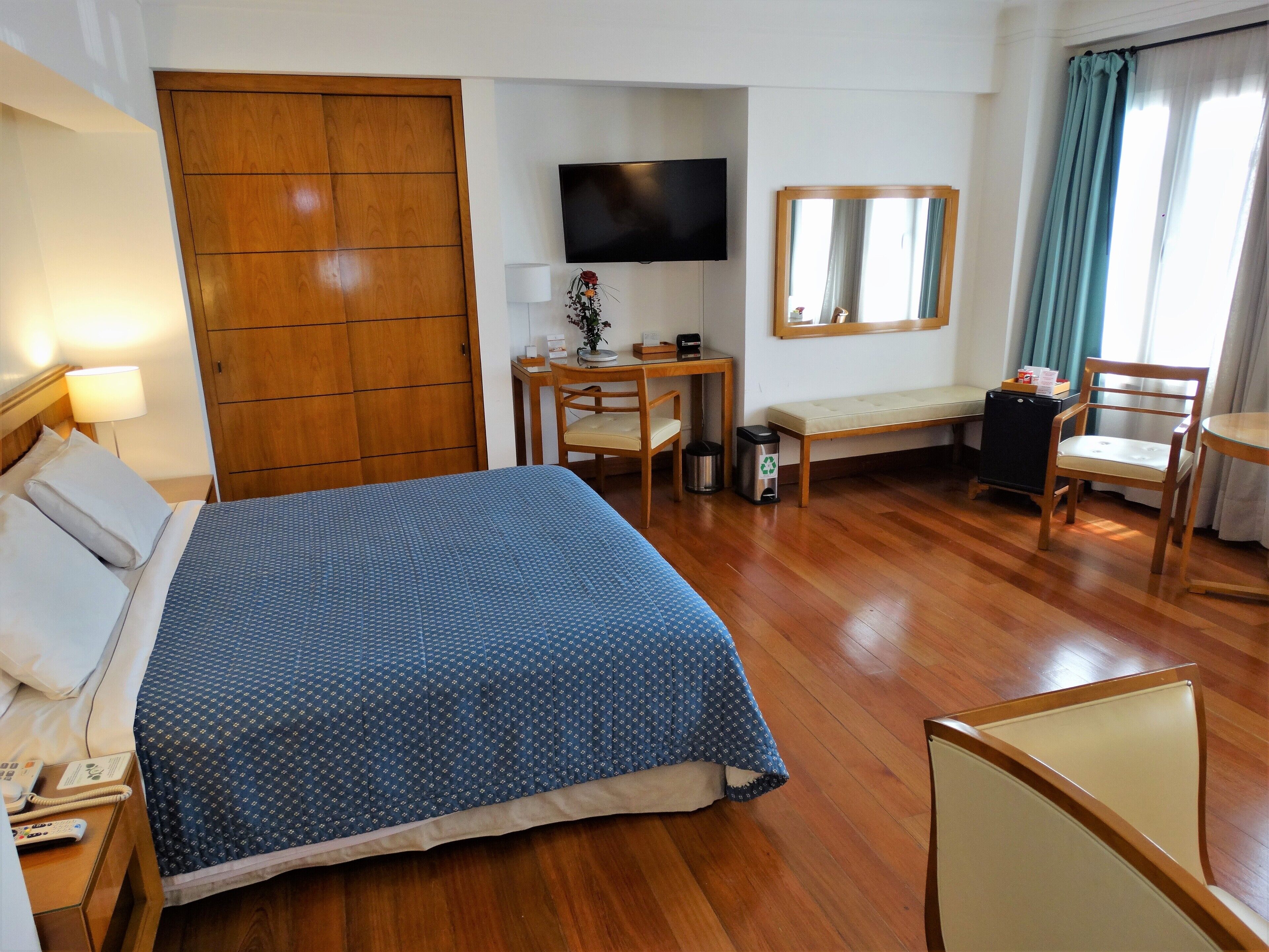 A hotel room with a bed, a desk, a chair, a TV, a mirror, and a window with curtains.