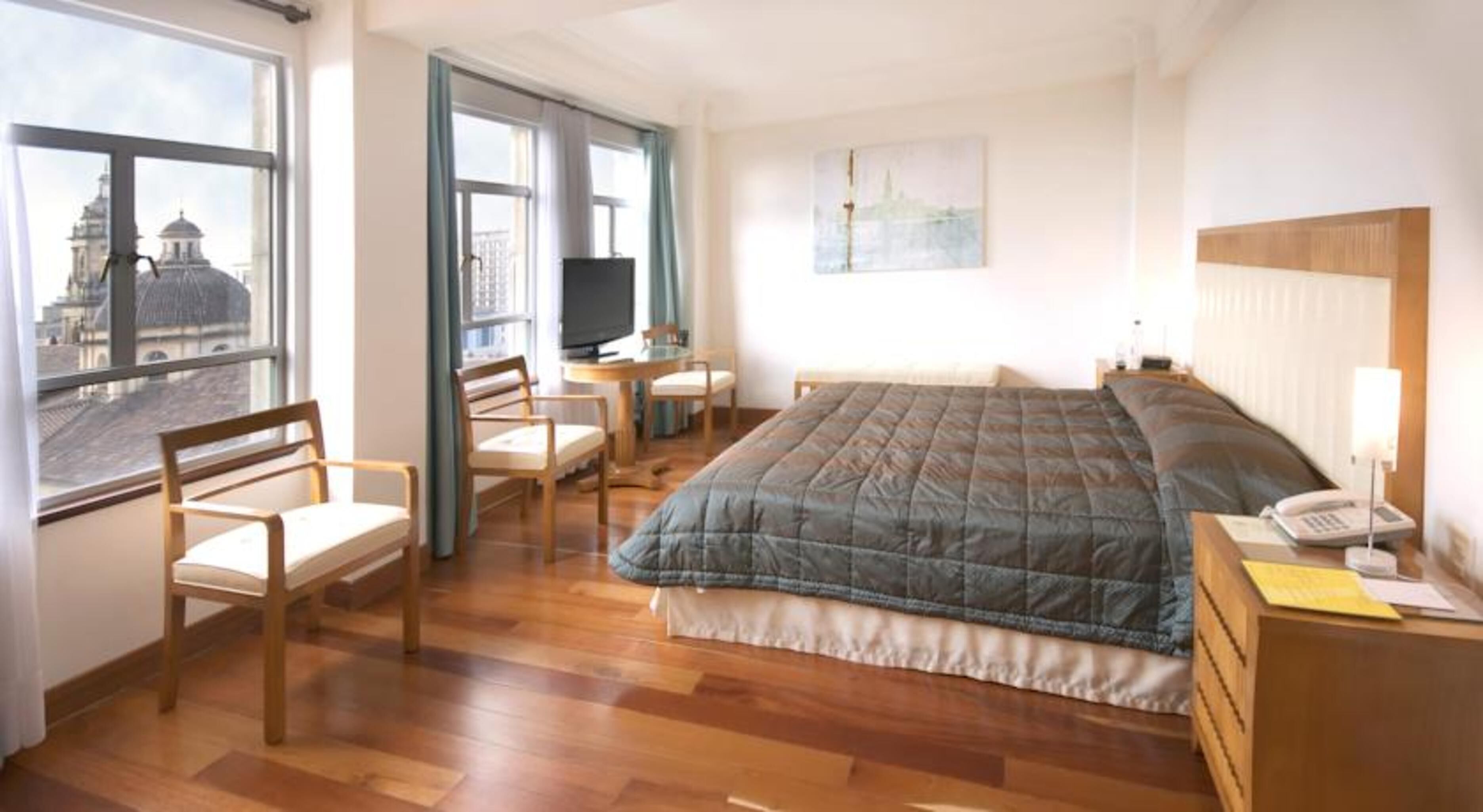 A hotel room with a bed, a desk, a chair, a TV, and a window with a city view.
