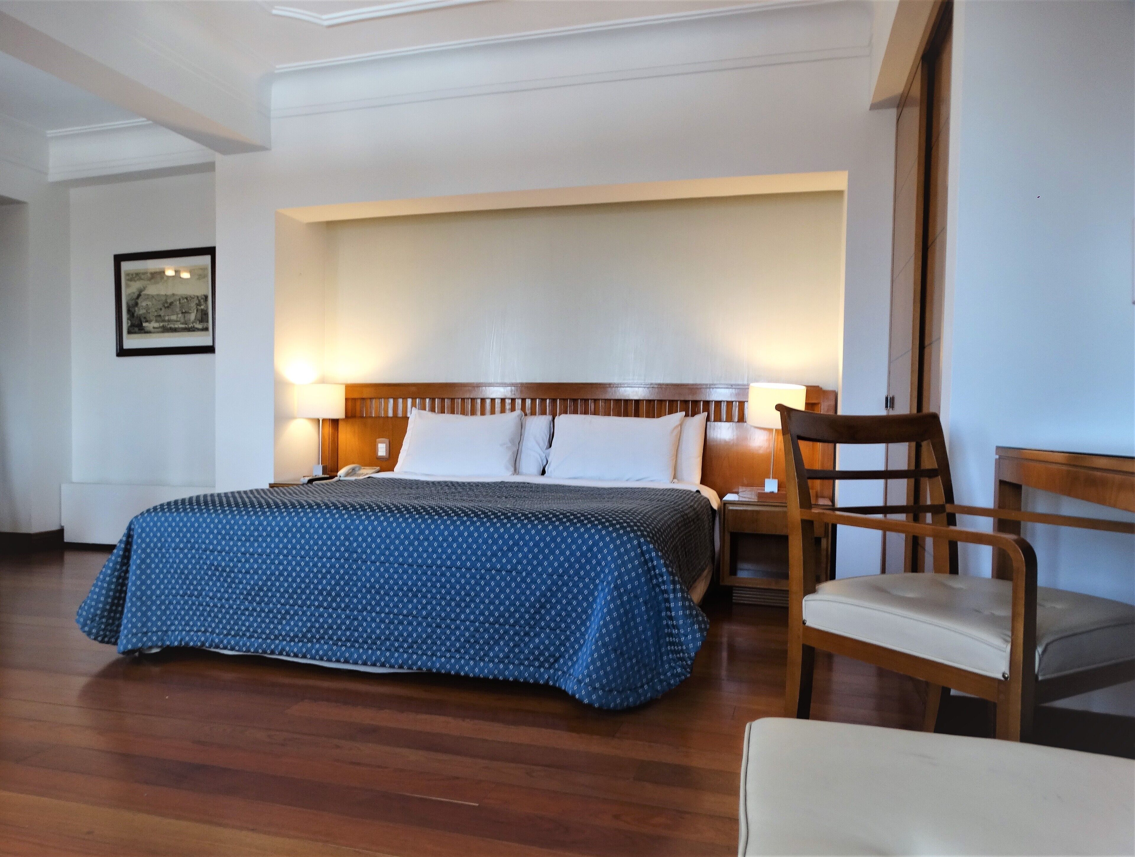 Quarto de hotel com uma cama grande, cabeceira de madeira, duas luminárias laterais, uma poltrona e um quadro na parede.