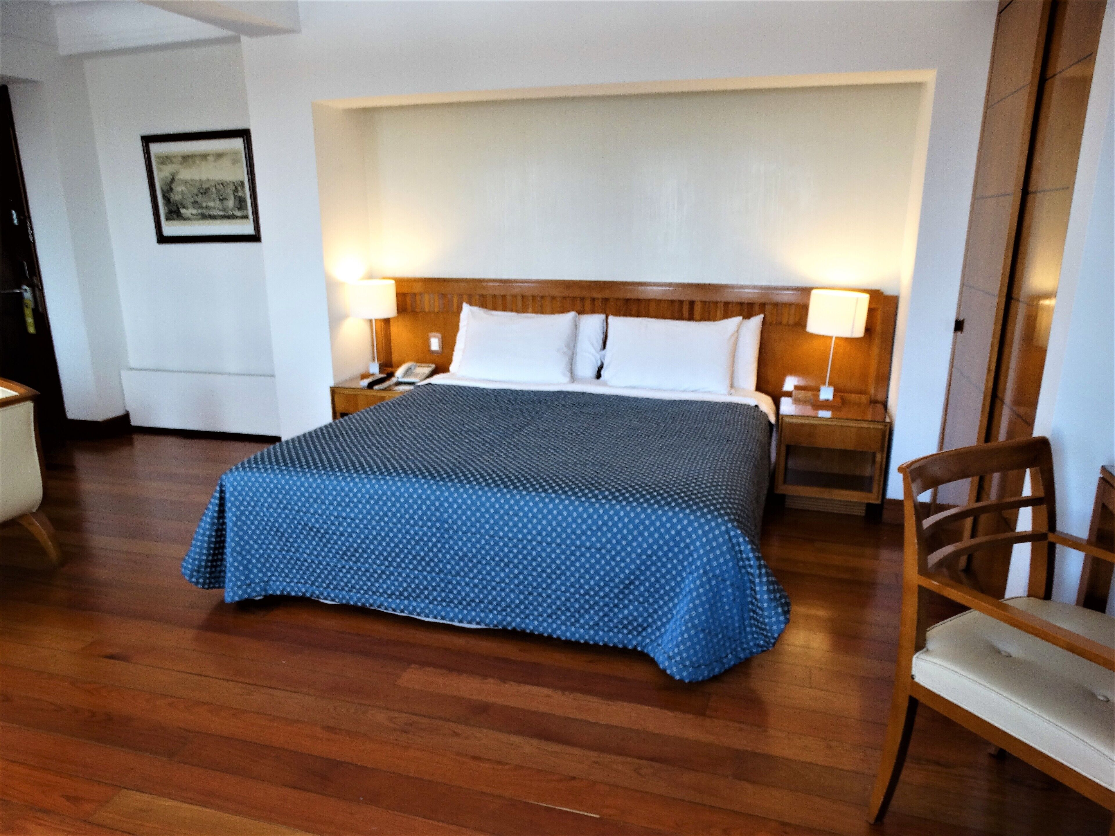 A hotel room with a large bed, wooden headboard, two bedside tables with lamps, a wooden chair, and a framed picture on the wall.