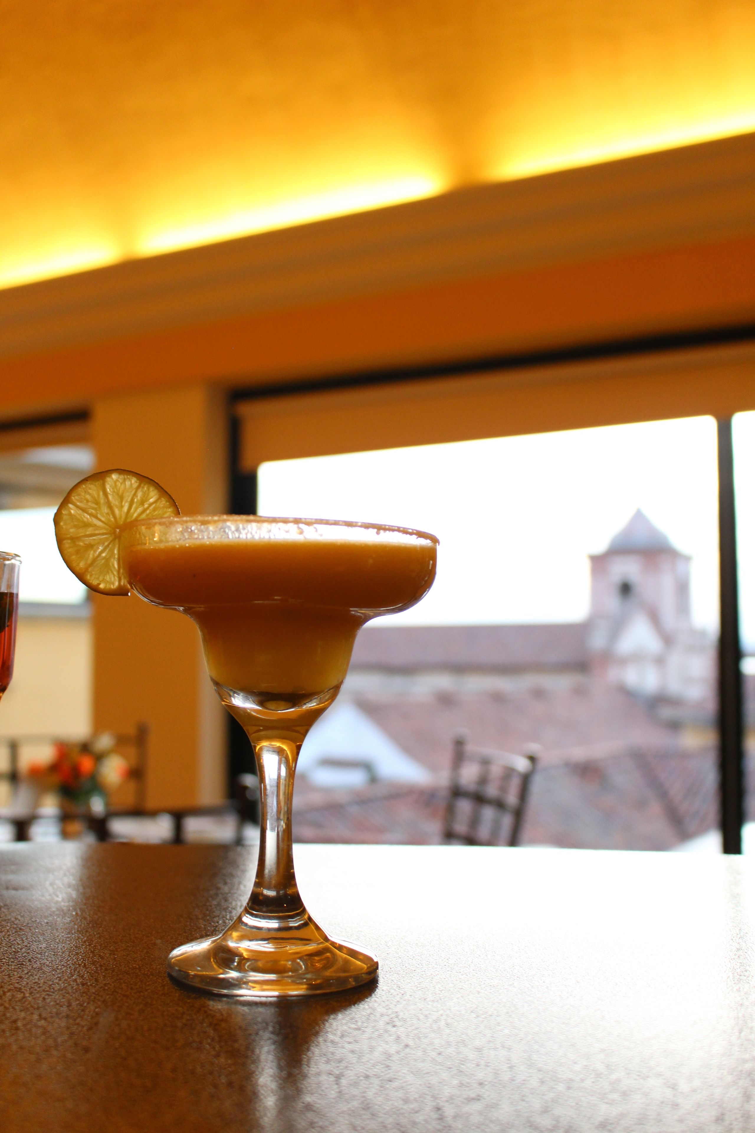 A cocktail glass with a slice of citrus on the rim, placed on a table with a view of a building in the background.
