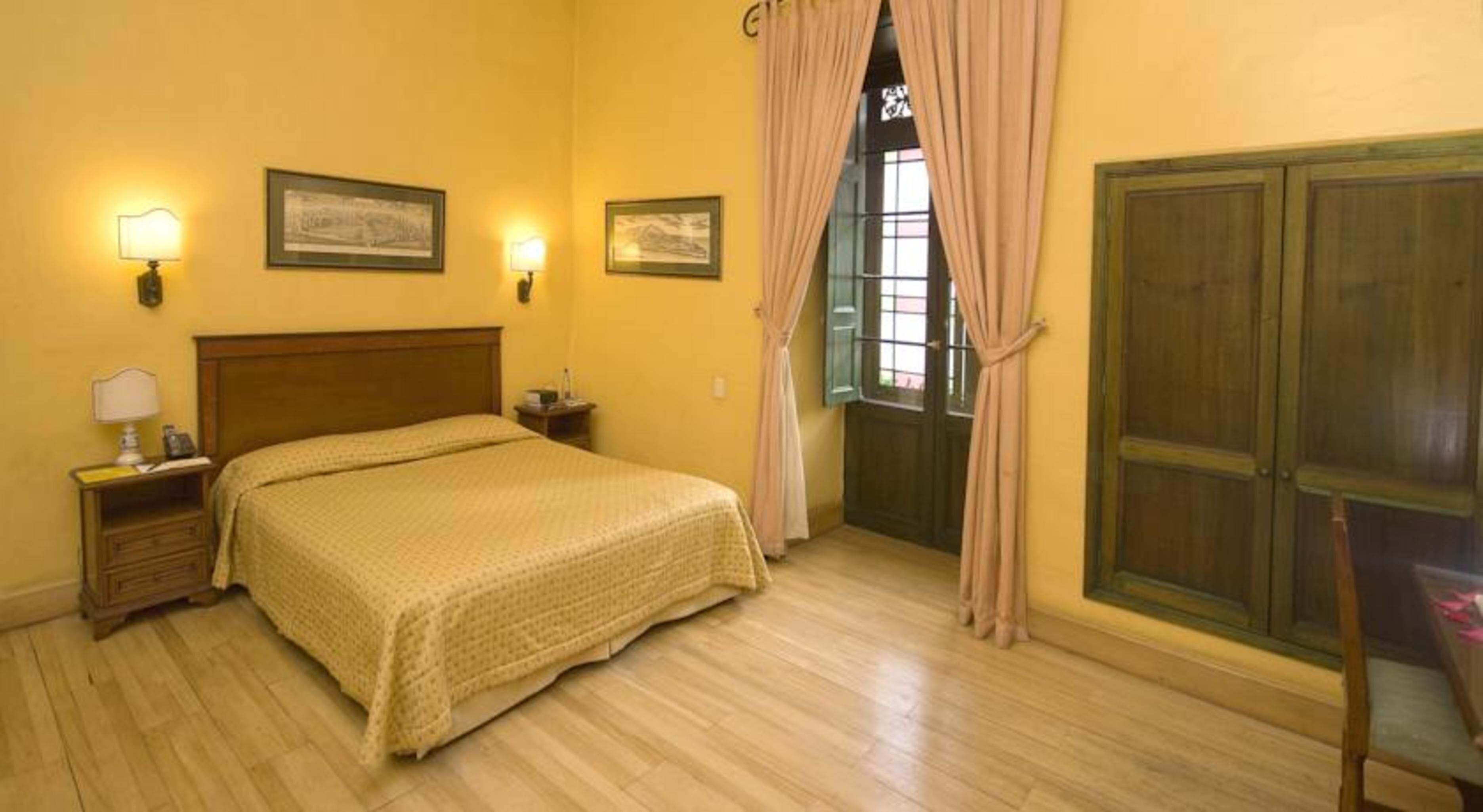 A bedroom with a wooden bed, bedside lamps, a nightstand, a window with curtains, and a wooden wardrobe.