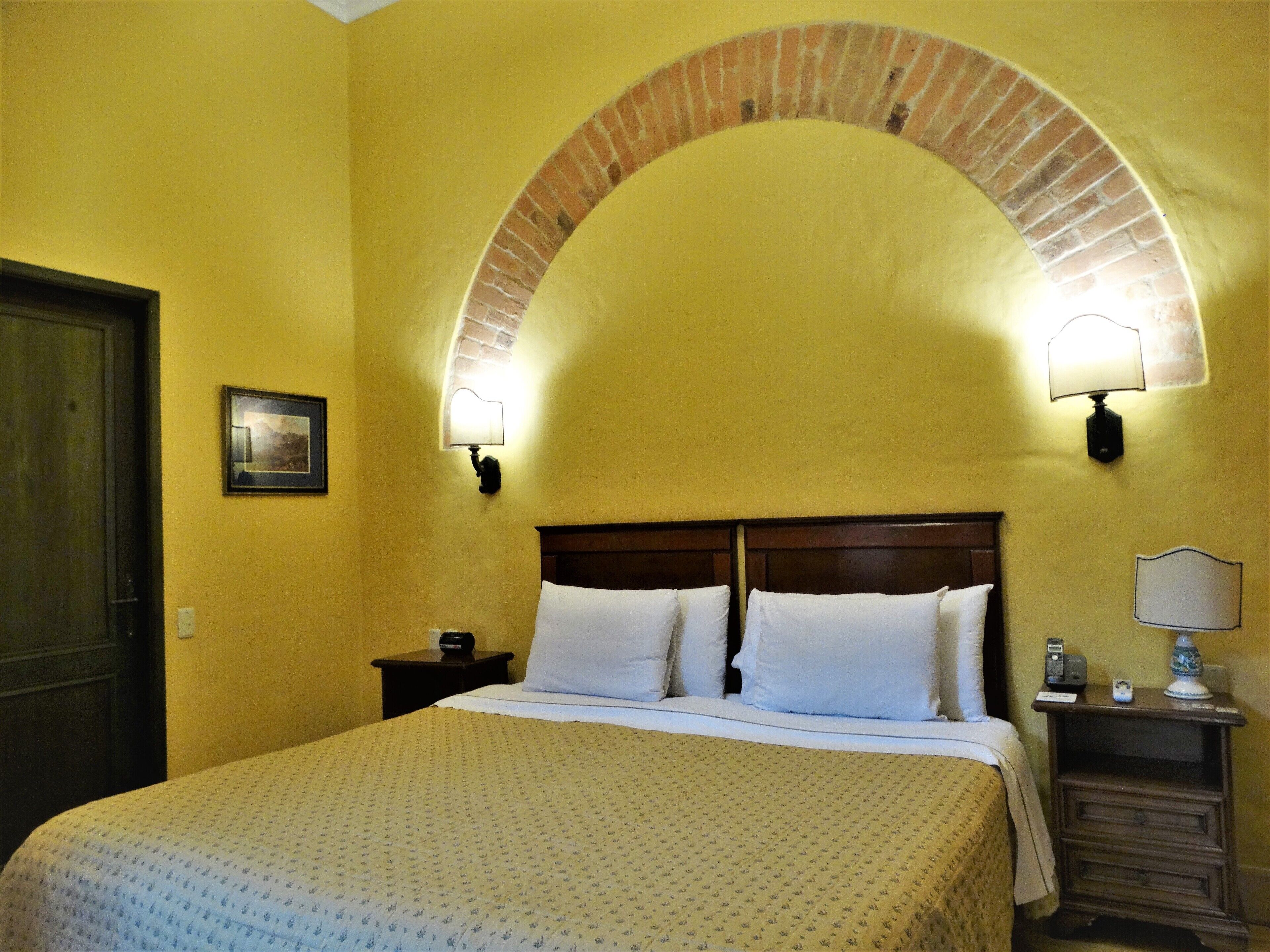 A hotel room with a bed, a nightstand, a lamp, and a brick archway.