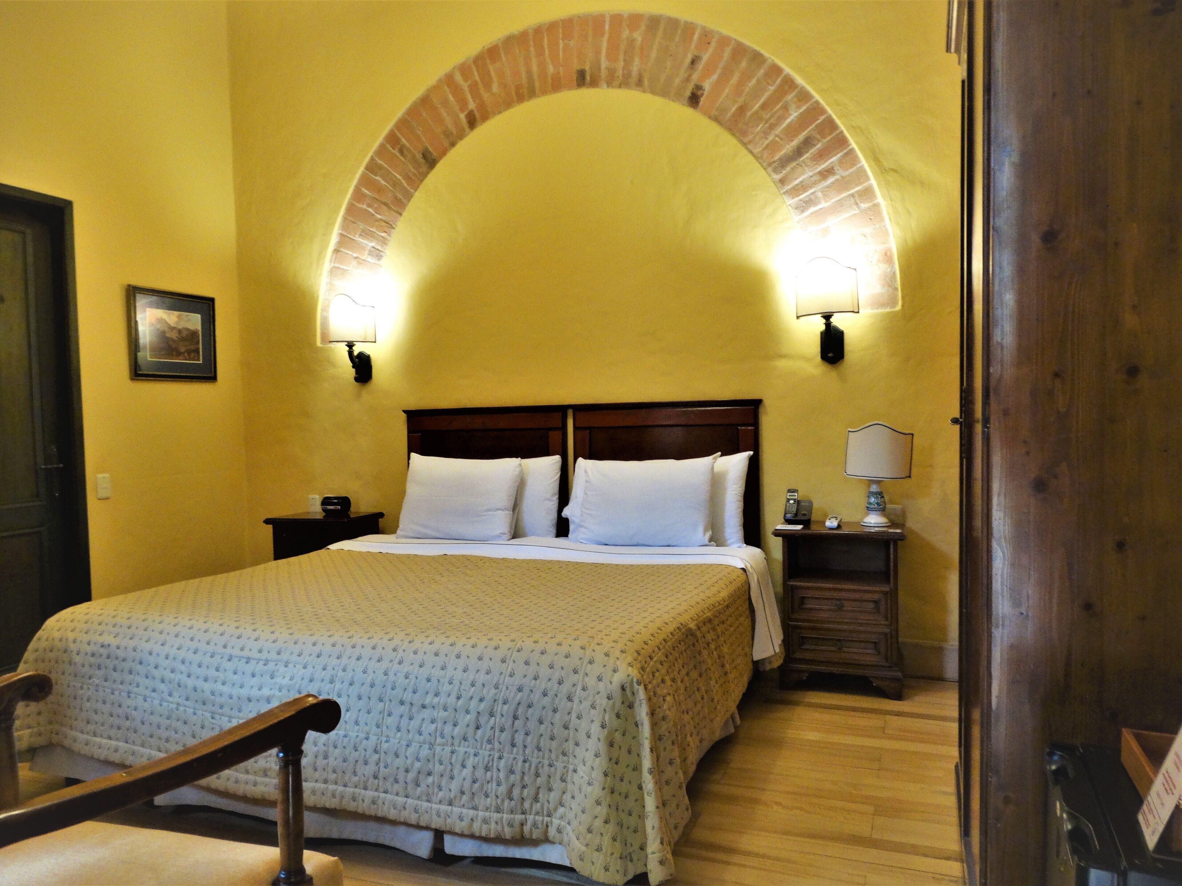 A hotel room with a bed, wooden headboard, two bedside tables with lamps, a picture on the wall, and an arched brick feature above the bed.