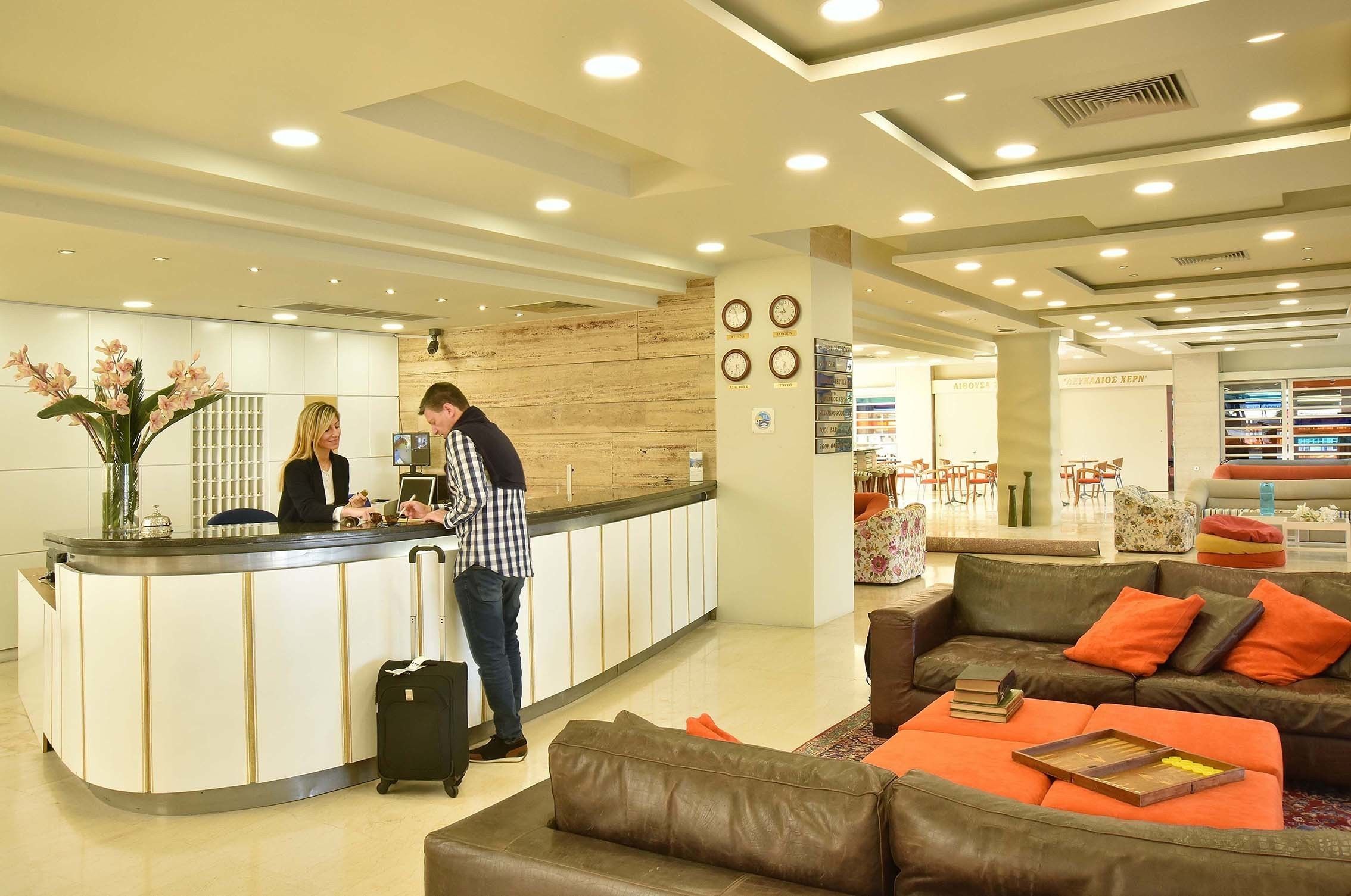 A hotel reception area with a counter, a waiting area with a sofa and armchairs, and a dining area in the background.