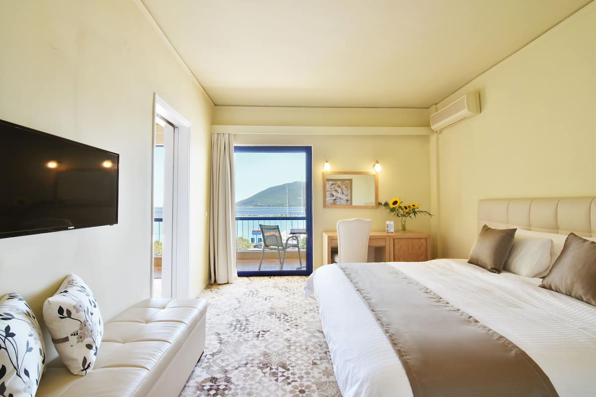 A hotel room with a large bed, a flat-screen TV mounted on the wall, a desk with a chair, and a balcony with a view of the sea.