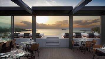 An outdoor dining area with tables and chairs, a view of the sea, and a sunset.