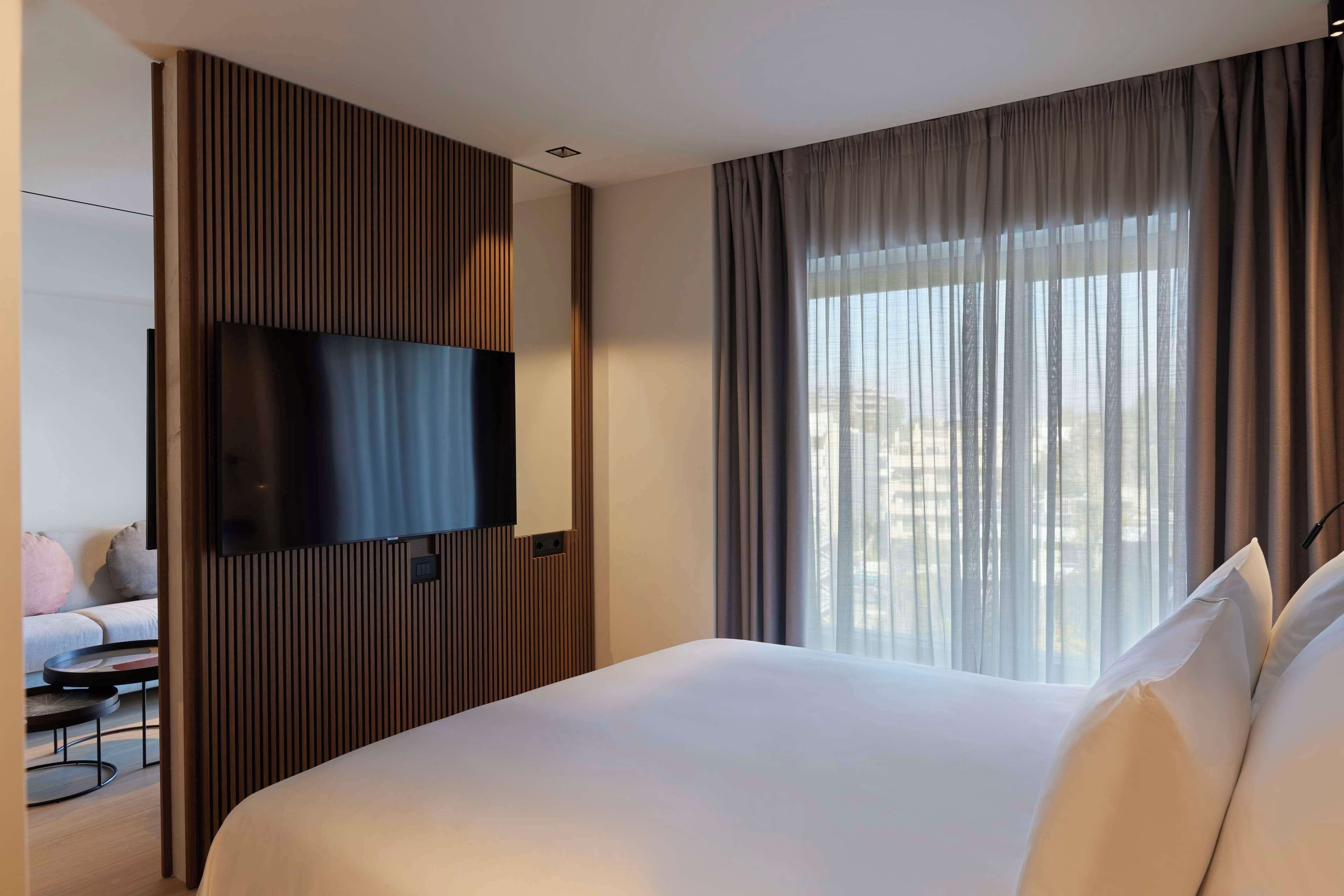 Ein modernes Hotelzimmer mit einem großen Bett, einem Flachbildfernseher an einer Wand aus Holzpaneelen und einem großen Fenster mit transparenten Vorhängen.
